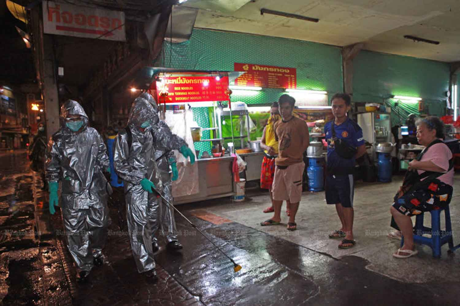 People watch as soldiers in full protective gear spray a disinfectant solution on the footpath in Bangkok's Yaowarat area in the early hours of Thursday. The army will be disinfecting various locations across the city until the end of the month to combat the spread of coronavirus. (Photo by Arnun Chonmahatrakool)