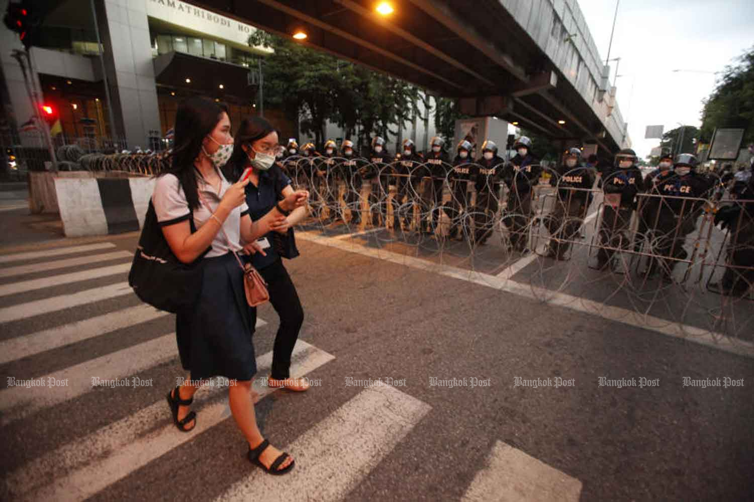 Two women in face masks walk past riot police blocking a road to Government House at Tuek Chai intersection, Bangkok, on Wednesday, when the country logged 10 new Covid-19 cases. (Photo: Pornprom Satrabhaya)