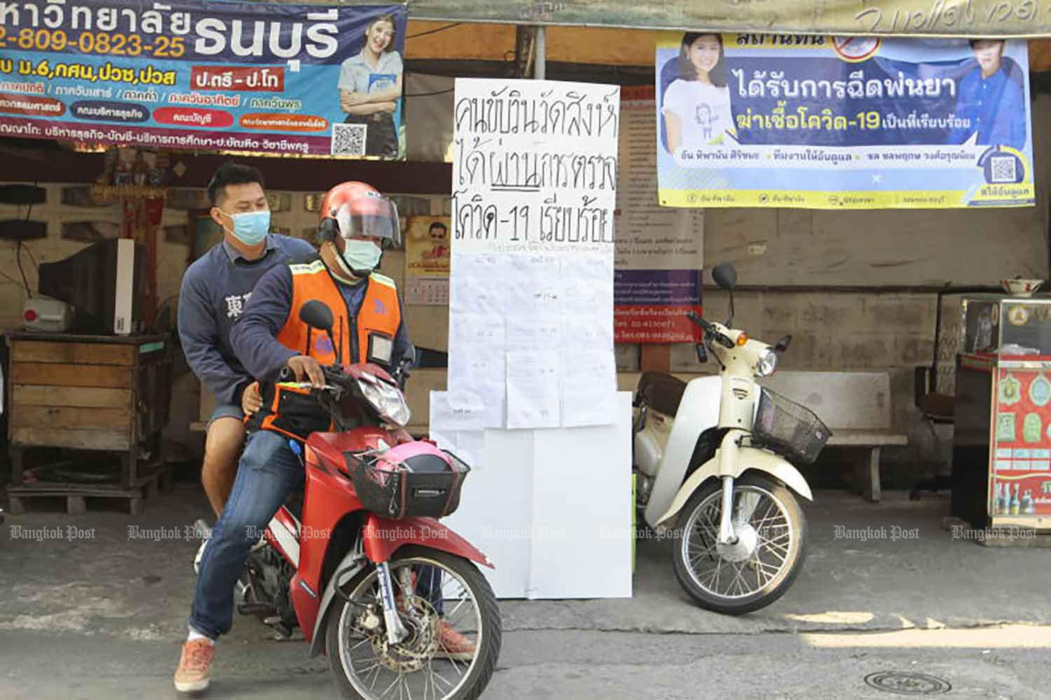 A taxi motorcyclist picks up a passenger on Soi Wat Sing in Chom Thong district of Bangkok on Tuesday, with a poster hung at the stand saying drivers there have been tested for Covid-19. (Photo: Arnun Chonmahatrakool)