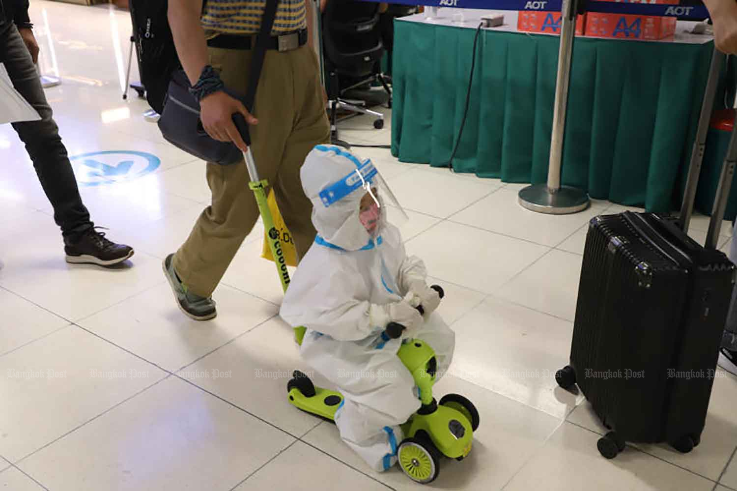 A child in full protective gear is pushed on a toddler's scooter as his family arrives at Suvarnabhumi airport, Samut Prakan province, on Monday. The Public Health Ministry will consider lowering the Covid-19 alert to Level 2 this week as cases continue to fall. (Photo: Varuth Hirunyatheb)