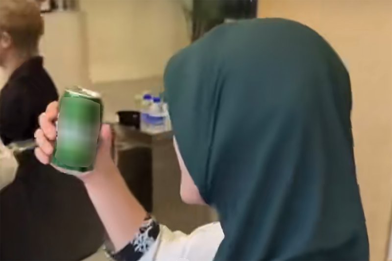 A screenshot from a video showing a man in a green headscarf drinking a can of beer at a hair salon in Malaysia.