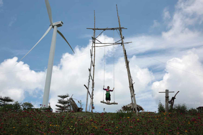 A person standing on a large wooden swing on a flower-covered hill next to a wind turbine.