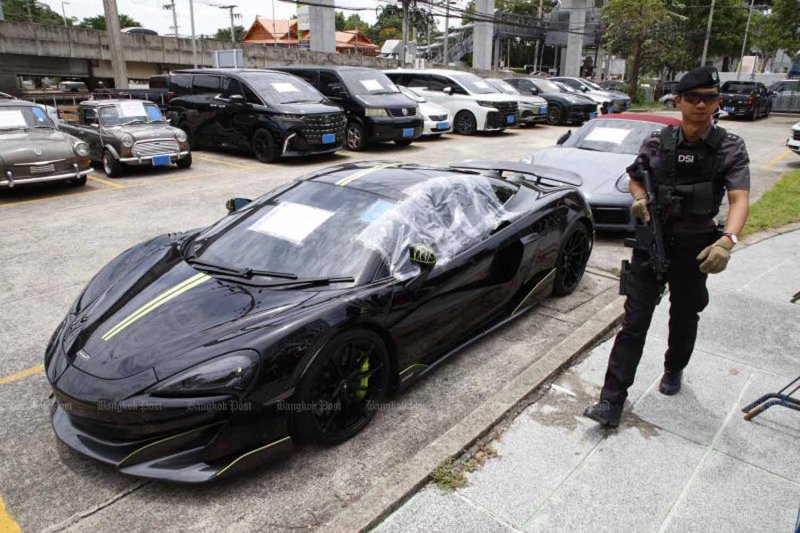 An armed DSI official walks past luxury cars at the agency’s headquarters in Bangkok on June 19. The vehicles were among assets worth 65 million baht impounded in the foreign-exchange scam case. (Photo: Pattarapong Chatpattarasill)