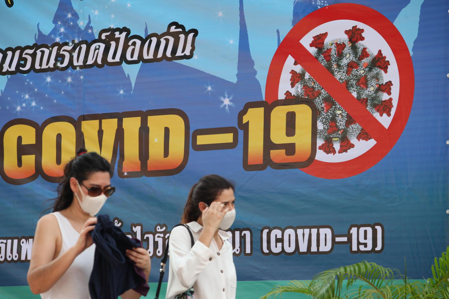 Women wearing face masks walk past a banner explaining how to prevent the spread of Covid-19. Apichart Jinakul