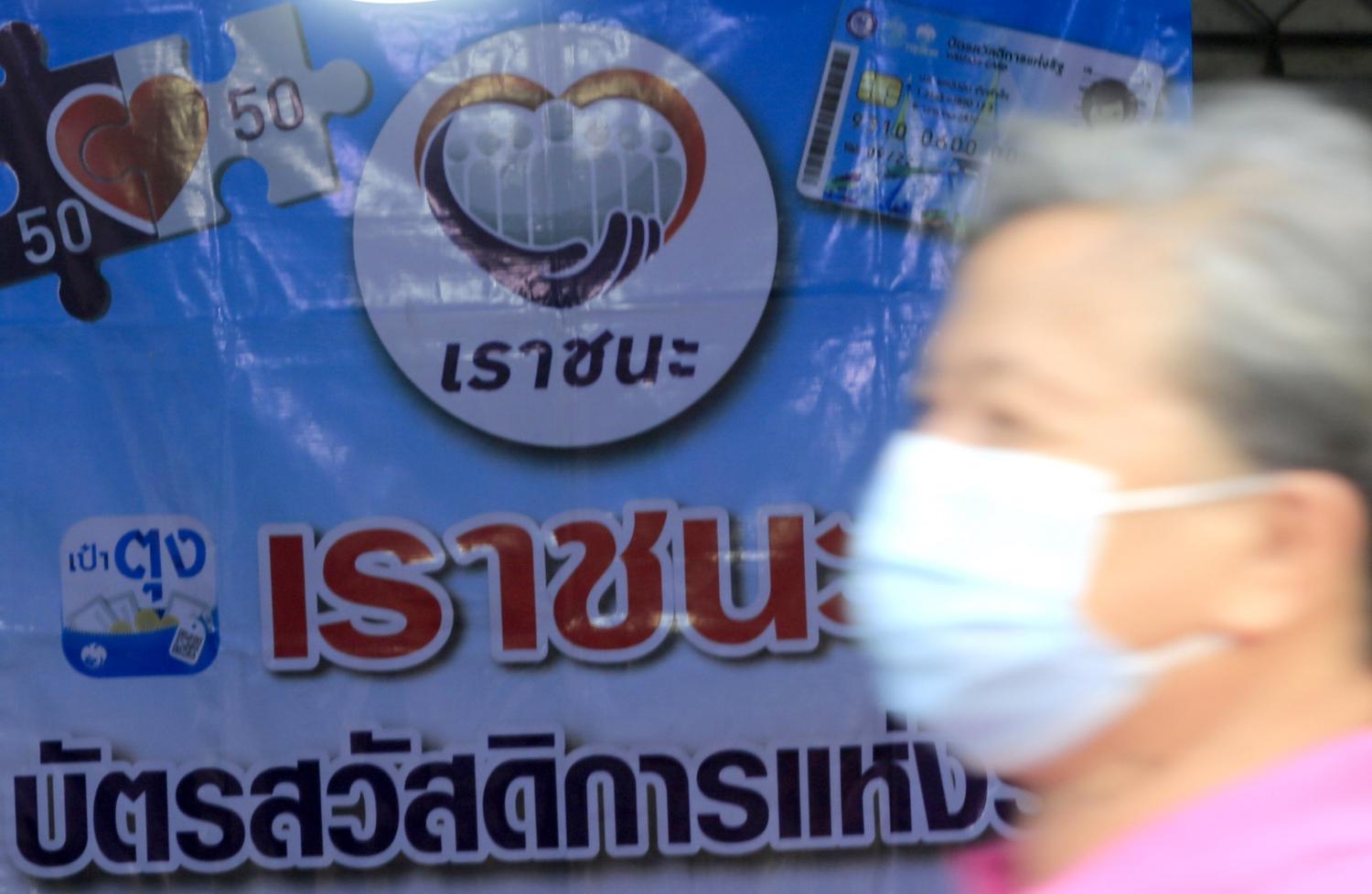 A man wearing a face mask walks past a sign of the Rao Chana (We Win) financial stimulus and the Kon La Krueng (Half-Half) co-payment scheme at Chatuchak weekend market. (Photo by Pornprom Satrabhaya)